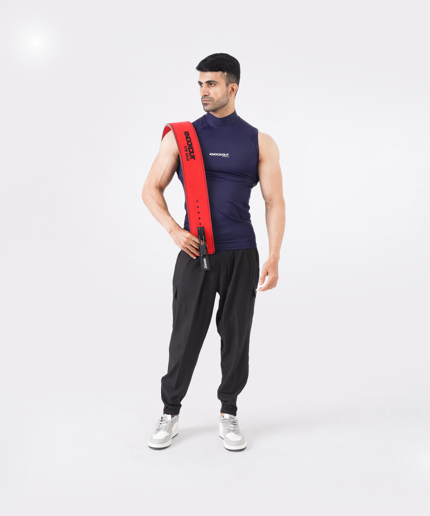 Man wearing a navy blue Knockout sleeveless high-neck compression shirt paired with black joggers, holding a red weightlifting belt.